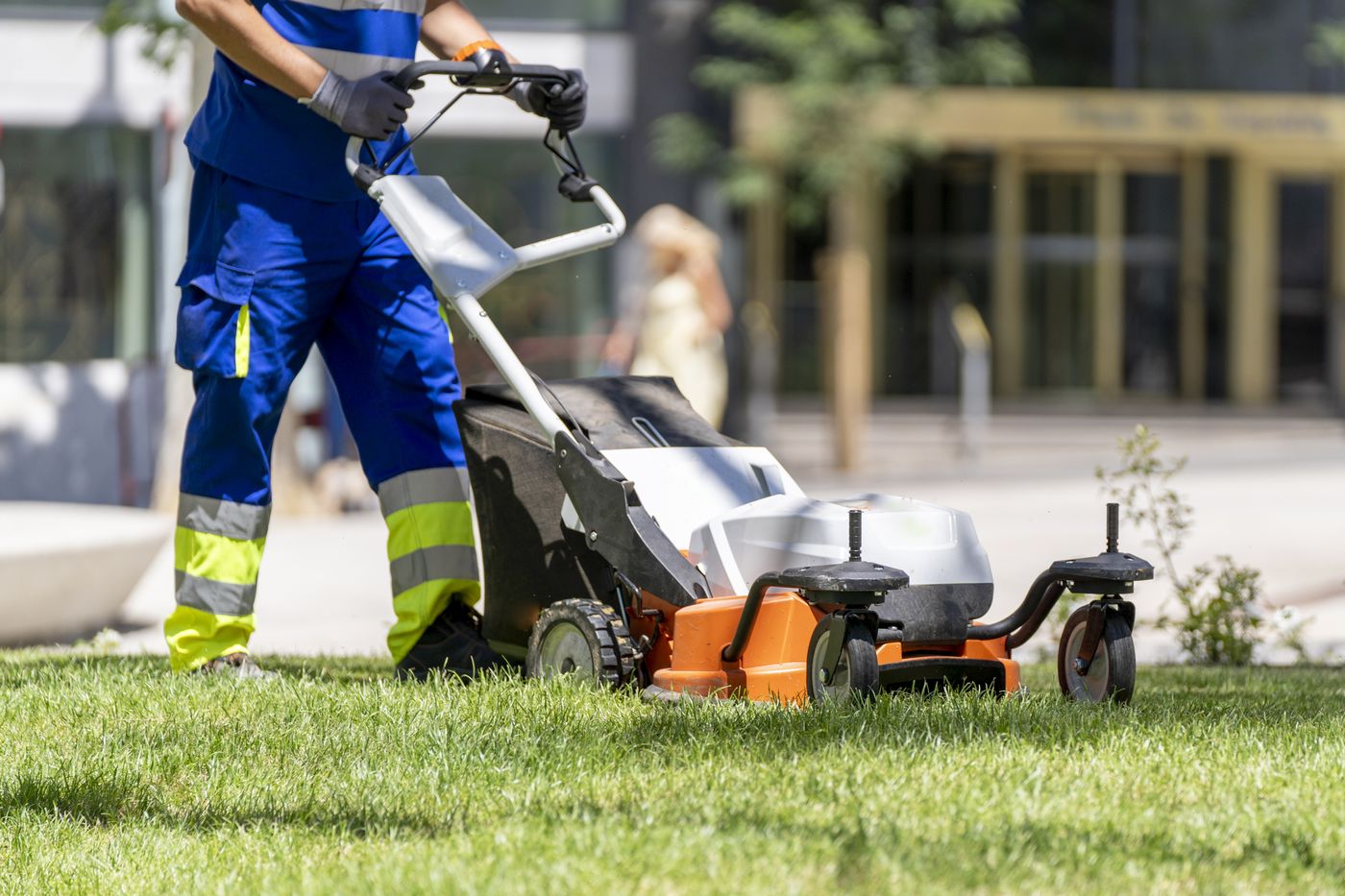 Worker in high-visibility trousers pushing a lawn mower across green grass